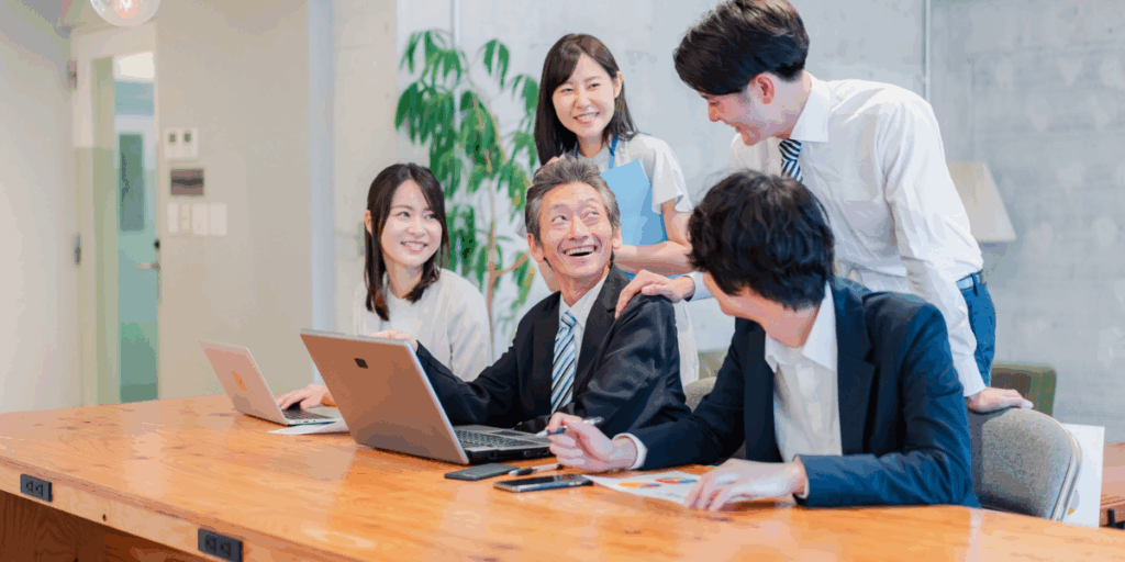 オフィスの会議テーブルで、ベテラン社員を中心に若手社員たちが笑顔で会話しながらパソコン作業をしている様子（世代を超えた職場のコミュニケーションイメージ）。